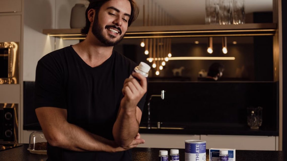 A smiling man in a kitchen holds a bottle of supplements, showcasing health and wellness.