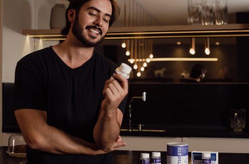 A smiling man in a kitchen holds a bottle of supplements, showcasing health and wellness.